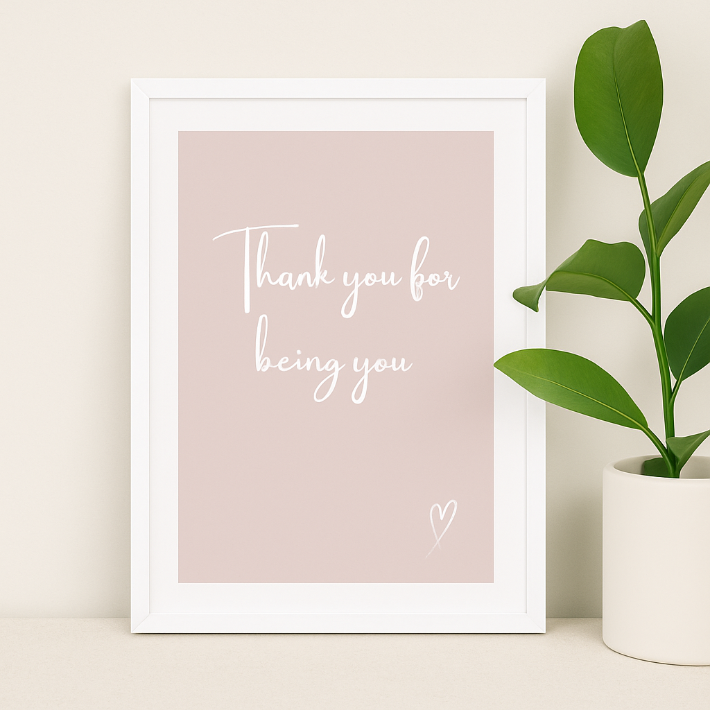 Framed print with 'Thank you for being you' text on a pink background, placed on a surface with a plant.