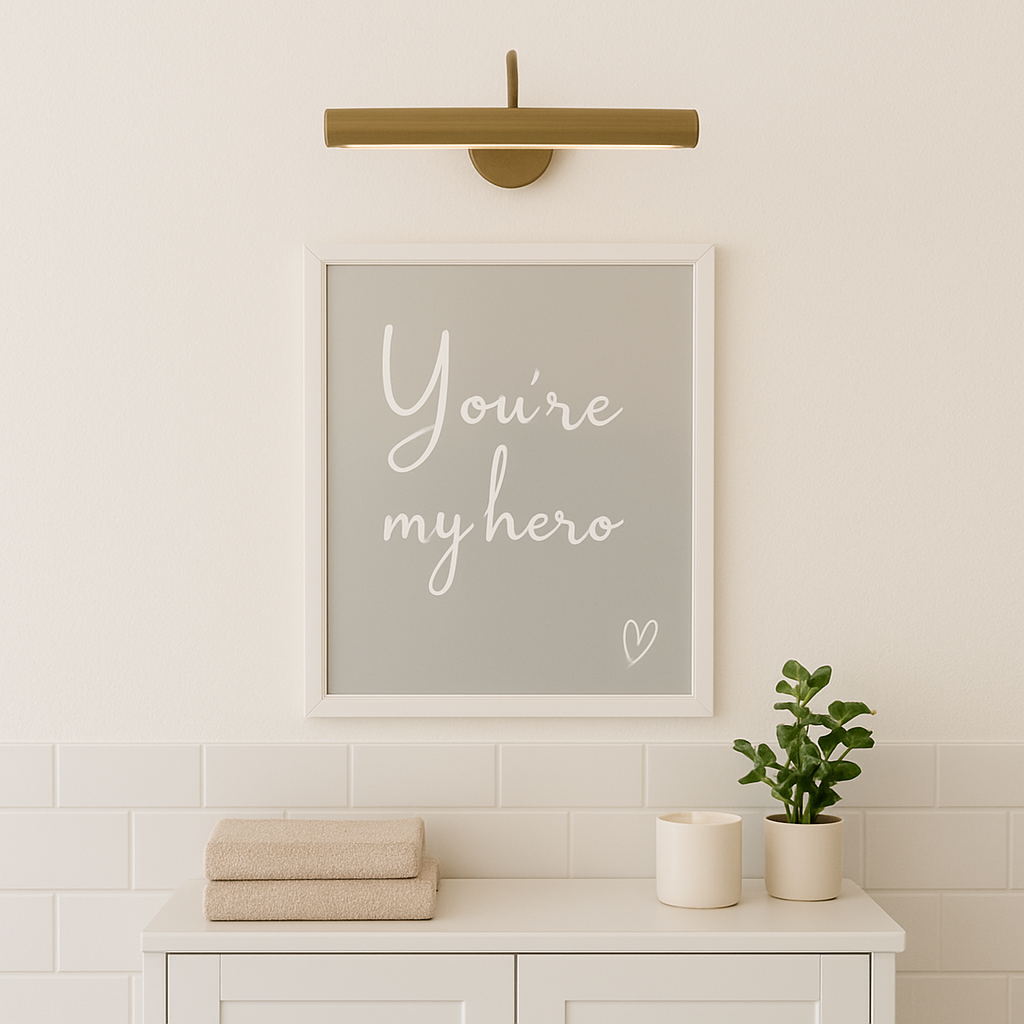 Framed artwork with 'You're my hero' text on a wall above a cabinet with towels and a plant.